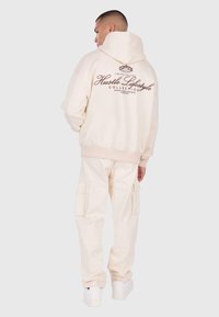 Mężczyzna w kremowych spodniach cargo i pasującej bluzie z kapturem z napisem "Hustle Lifestyle Collection" na plecach, stojący na jednolitym tle.
