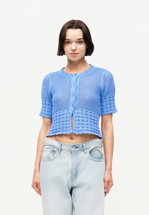 Giovane donna con capelli corti e scuri che indossa un cardigan blu corto a maniche corte lavorato a maglia e jeans chiari a vita alta su uno sfondo semplice.