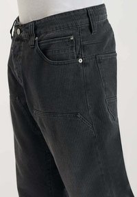 Pantalon en velours côtelé noir avec une texture côtelée, doté de deux poches avant et d'un bouton brillant. La taille est élastiquée pour le confort.