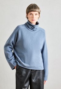 Svetlo modra turtleneck pulover iz mehkega pletenega materiala, s sproščenim krojem in rebrastimi manšetami, v kombinaciji z črnimi usnjenimi hlačami.