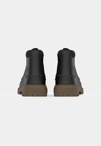 Botas de cuero negras hasta el tobillo con un acabado liso, cuello acolchado y suela de goma marrón con dibujo, presentando un diseño minimalista.
