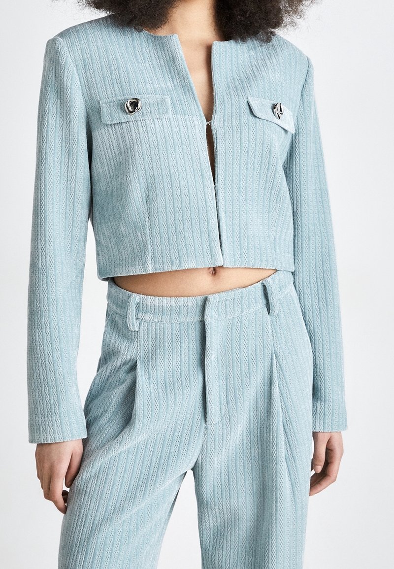 Femme portant une veste courte texturée bleu clair avec des boutons argentés et un pantalon taille haute assorti sur fond blanc.