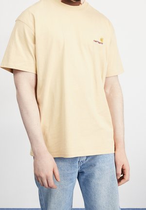 Hellgelbes Baumwoll-T-Shirt mit entspannter Passform, kurzen Ärmeln und einem kleinen roten gestickten Logo auf der linken Brustseite. Kombiniert mit hellblauen Jeans.