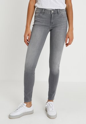 Personne portant un jean slim gris et des baskets blanches, debout devant un fond clair uni.