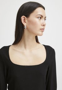 Schwarzes, geripptes Oberteil mit quadratischem Ausschnitt und gewelltem Saum; das Model hat glattes, dunkles Haar und trägt silberne Creolen.