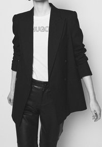 Blazer negro de corte oversized con botones de doble botonadura, usado sobre una camiseta blanca con letras negras. Los pantalones de cuero negro ajustados completan el conjunto.