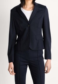 Blazer bleu marine en tissu lisse, doté d'un col à revers cranté, d'une fermeture à un bouton et de deux poches avant, avec une silhouette ajustée.
