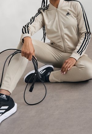 Personne en survêtement beige Adidas et baskets noires Adidas assise en tailleur tenant une corde à sauter noire sur un sol gris.