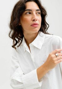 Hvid skjorte med knapper, dekoreret med små sølvprikker, lange ærmer og krave; modellen bærer en sølvring på sin venstre hånd.