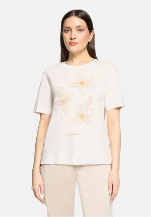 Femme portant une chemise blanche à manches courtes avec trois fleurs beiges brodées et le texte « FLOWER GARDEN », associée à un pantalon beige.