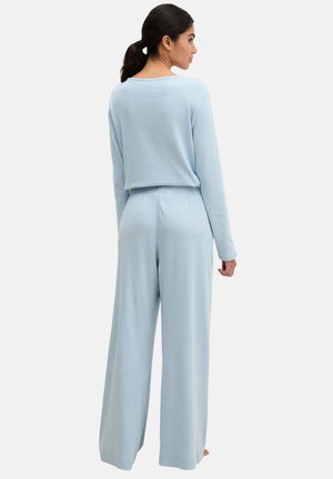 Kobieta o ciemnych włosach związanych w kucyk, ubrana w jasnoniebieski prążkowany top z długim rękawem i szerokie spodnie, stoi tyłem.