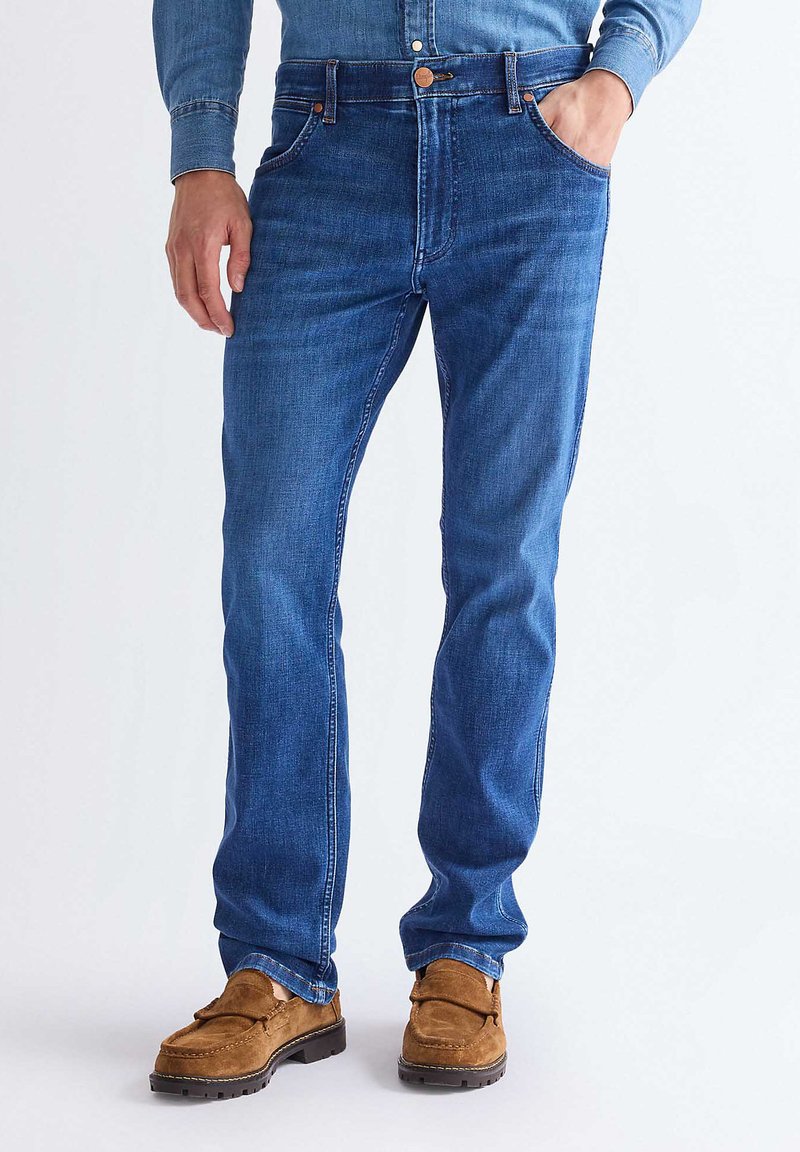 Mann iført blå jeans, brune mokasiner i semsket skinn og en blå skjorte med knapper, med den ene hånden i lomma, mot ensfarget bakgrunn.