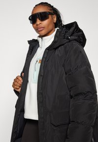 Schwarze gepolsterte Parkajacke mit Kapuze, mit einem Reißverschluss und einem mittellangen Design. Begleitet von einem weißen Fleece und übergroßen Sonnenbrillen.