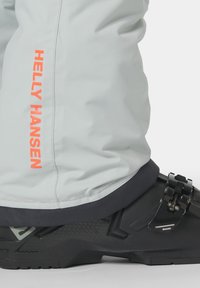 Světlé šedé lyžařské kalhoty s oranžovým logem "Helly Hansen"; mají černý nastavitelný botník s texturovaným povrchem a kovovými akcenty.