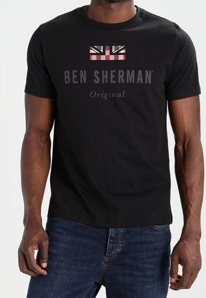 T-shirt noir Ben Sherman avec un Union Jack stylisé et le texte "BEN SHERMAN Original", porté par une personne en jean bleu.