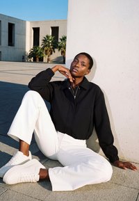 Vrouw in een zwart jasje, witte broek en witte sneakers die buiten tegen een witte muur zit met palmbomen en gebouwen op de achtergrond.