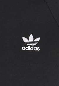 Tessuto nero con una finitura testurizzata, caratterizzato da un logo Adidas ricamato in bianco e un design stilizzato a tre foglie sopra il nome del marchio.