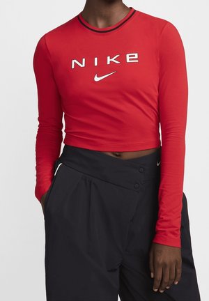 Osoba ubrana w czerwony krótki top z długim rękawem z białym logo Nike oraz czarne spodnie z wysokim stanem, ręce swobodnie ułożone wzdłuż ciała.