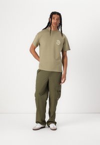 Olive green cotton t-shirt with a circular white graphic design on the chest, paired with olive cargo pants featuring side pockets.