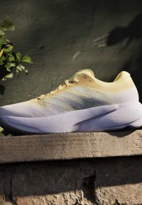 Ανοιχτό κίτρινο και λευκό αθλητικό παπούτσι με τρεις πλευρικές ρίγες, τοποθετημένο σε τραχιά πέτρινη επιφάνεια με σκοτεινό, θολό φόντο.
