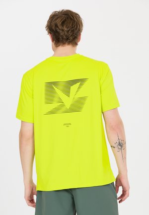 Mand iført lysegul T-shirt med sort abstrakt logo på ryggen, viser tatovering på højre underarm, stående mod en hvid baggrund.