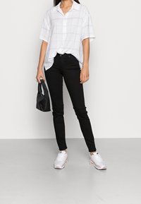 Camisa blanca de cuadros con mangas cortas, jeans negros ajustados y acentos rosas en las zapatillas. Bolso negro en la mano, de pie sobre una superficie clara.