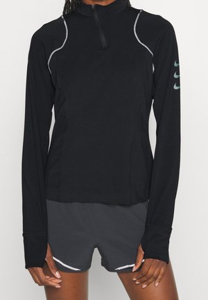 Person wearing a black long-sleeve athletic top with zipper and logos on sleeve, paired with black athletic shorts with reflective details.