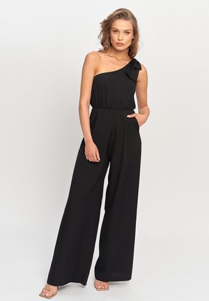 Kobieta ubrana w czarny kombinezon na jedno ramię z szerokimi nogawkami i detalem w formie kokardki na ramiączku, stoi z jedną ręką w kieszeni.