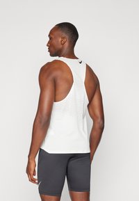 Weißes sportliches Tanktop mit Netzdesign, im Racerback-Schnitt und einem kleinen schwarzen Logo auf dem oberen Rücken, kombiniert mit schwarzen Shorts.