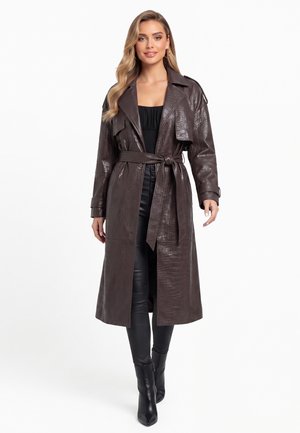 Vrouw draagt een lange donkerbruine, getextureerde trenchcoat met ceintuur, gecombineerd met een zwarte broek en zwarte enkellaarzen, tegen een witte achtergrond.