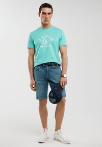 Minta zaļa T-krekls īsām piedurknēm ar "DENIM" grafiku, zilas džinsu šorti un baltas augstās kedas. Rokās tur zilu cepuri. Vienkāršs dizains.