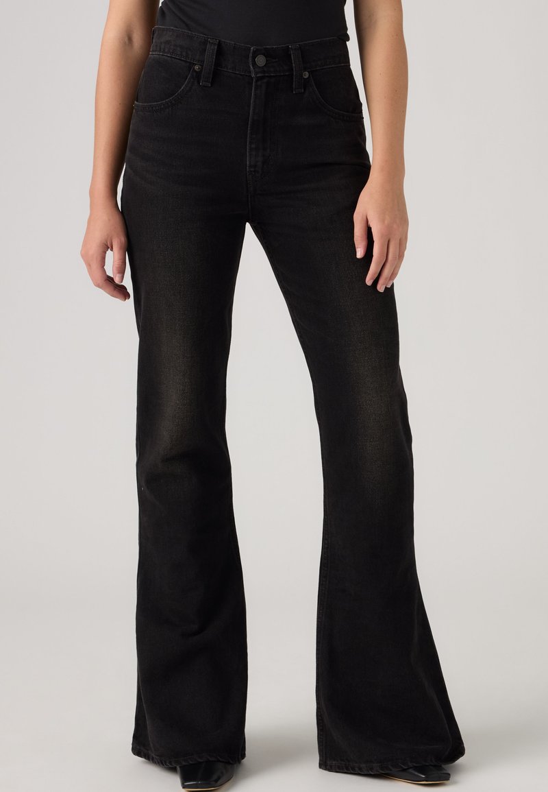 Femme portant un jean noir taille haute évasé et un haut noir, debout, les mains détendues le long du corps, sur un fond uni.