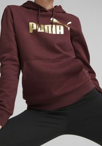 Felpa bordeaux con logo "PUMA" dorato. Presenta una tasca a canguro, cappuccio con cordoncino e polsini e orlo a coste. Tessuto dalla texture liscia.