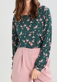 Donkergroene blouse met lange mouwen, voorzien van een bloemenpatroon in roze. Combinatie met lichtroze high-waisted broek.