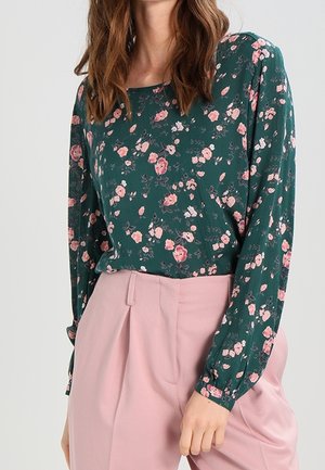Dunkelgrüne Bluse mit langen Ärmeln, verziert mit einem floralen Muster in Rosa. Kombiniert mit hellrosa, hoch taillierten Hosen.