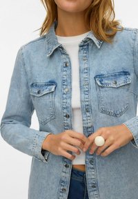 Camicia in denim azzurro chiaro con chiusura a bottoni, due tasche sul petto e accessori in contrasto. Il modello indossa un anello bianco e una t-shirt sotto.