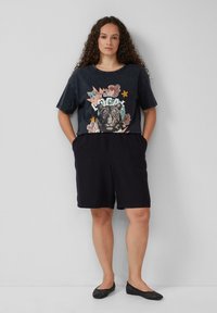 Donkergrijze graphic t-shirt met een tijgerprint en kleurrijke accenten, gecombineerd met zwarte shorts en zwarte instap-schoenen met studs.