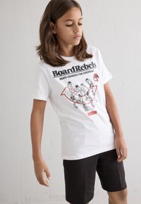 Hvit bomulls t-skjorte med grafisk trykk i svart og rødt, sammen med svarte shorts. Enkel design, kortermet, avslappet passform.