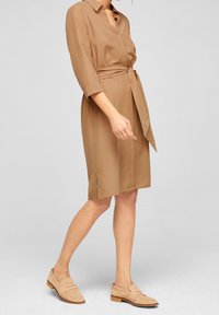 Abito camicia marrone con cintura, maniche a tre quarti, colletto e spacchi laterali, abbinato a mocassini marroni chiari.