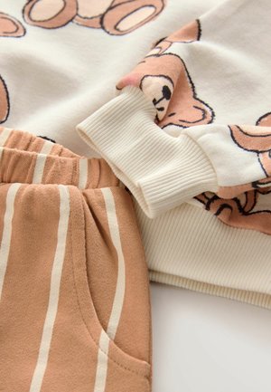 Weißer Pullover mit Bärenmotiv, gerippte Bündchen, kombiniert mit hellbraunen gestreiften Hosen mit Seitentasche und weicher Textur.