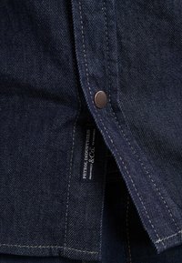 Camisa de denim azul oscuro con un acabado texturizado, que presenta un botón de metal y una etiqueta con la marca cosida en el lado inferior.