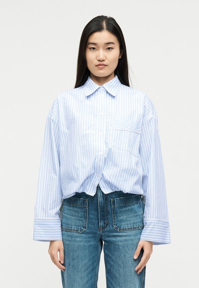 Νεαρή γυναίκα με μακριά σκούρα μαλλιά, φορώντας ένα φαρδύ μπλε και λευκό πουκάμισο με ρίγες και μπλε τζιν με μεγάλες μπροστινές τσέπες.