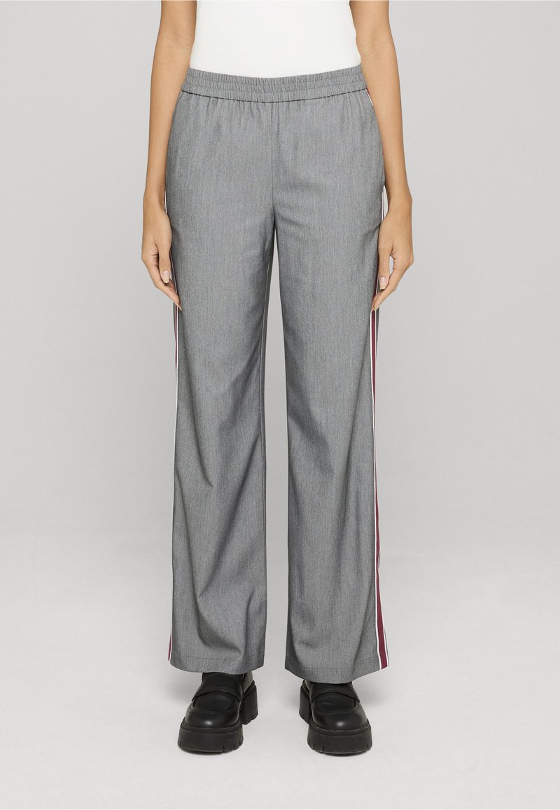 Pantalon large gris avec taille élastique et bandes latérales bordeaux et blanches, porté avec des chaussures noires épaisses sur une personne debout.