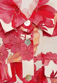 Koszulka w kwiaty z czerwonymi i różowymi wzorami, z zapięciem na guziki z przodu i kołnierzykiem. Materiał jest lekki o fakturowanym wyglądzie.