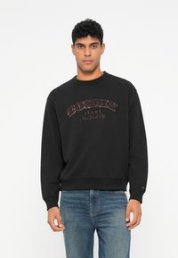 Čierna mikina s okrúhlym výstrihom a rebrovanými manžetami, ktorá má veľké zdobné logo "TOMMY JEANS New York City" v kontrastných farbách.