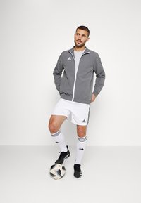 Graue Sportjacke mit weißem Reißverschluss, weißes T-Shirt, weiße Shorts mit schwarzen Streifen, schwarze Fußballschuhe und ein gemusterter Fußball.