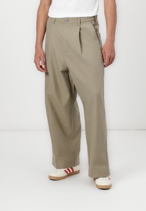 Pantalons plissés en kaki avec une coupe décontractée, fabriqués en tissu robuste. Comprennent un bouton avant et des détails de poches, complétés par des chaussures blanches à la cheville.