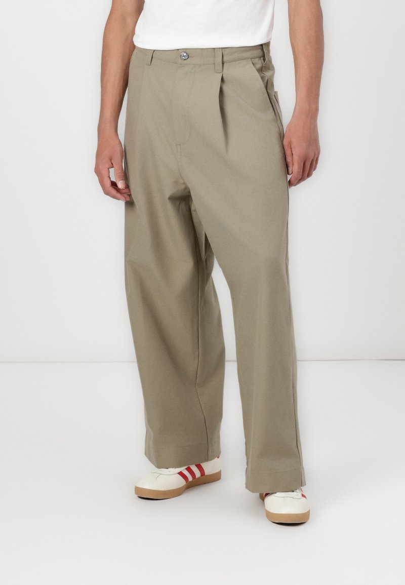 Khaki pleated trousers with a relaxed fit, made of sturdy fabric. Contains a front button and pocket details, complemented by white ankle shoes.