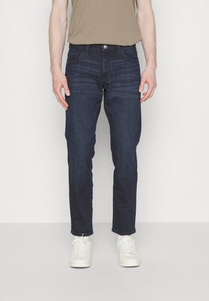 Donkerblauwe denim jeans met een reguliere pasvorm, vijf zakken en een knoopsluiting. Gecombineerd met witte sneakers, rustend op een lichtgrijze achtergrond.