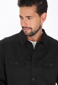 Camicia nera a bottoni con colletto, due tasche sul petto e un tessuto liscio e testurizzato. Abbinata a una maglietta bianca sottostante.